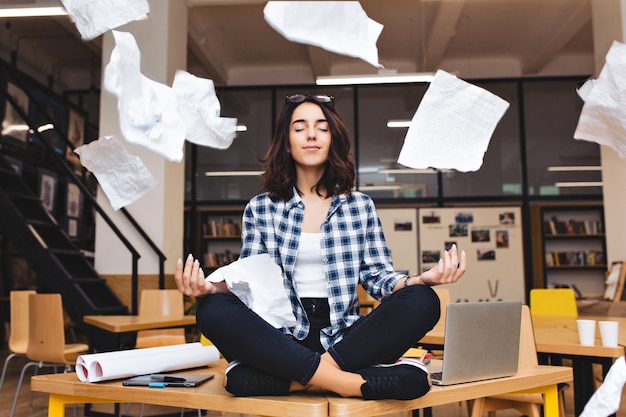 Jovem mulher morena muito alegre meditando sobre a mesa, cercar as coisas de trabalho e papéis voando. humor alegre, pausa, trabalho, estudo, relaxamento, verdadeiras emoções.