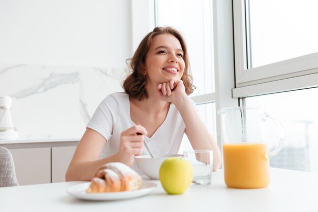 jovem mulher morena sorridente em camiseta branca tomando café da manhã saudável enquanto localização na mesa da cozinha