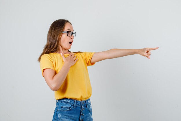 Jovem mulher mostrando algo em t-shirt, shorts, óculos e parecendo chocada