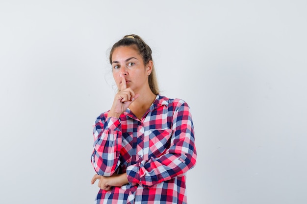 Jovem mulher mostrando o gesto de silêncio na camisa casual e olhando sensata, vista frontal.