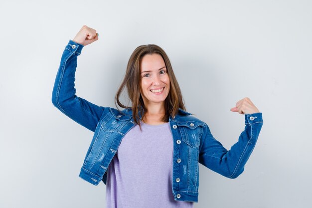 Jovem mulher mostrando o gesto do vencedor em t-shirt, jaqueta e parecendo com sorte, vista frontal.