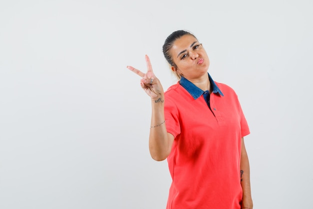 Jovem mulher mostrando o símbolo da paz, curvando os lábios em uma camiseta vermelha e bonita, vista frontal.