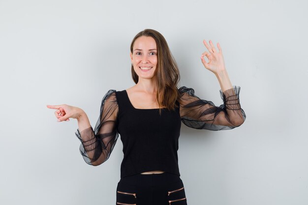 Jovem mulher mostrando sinal de ok e apontando para a esquerda em blusa preta e calça preta e parecendo feliz, vista frontal.