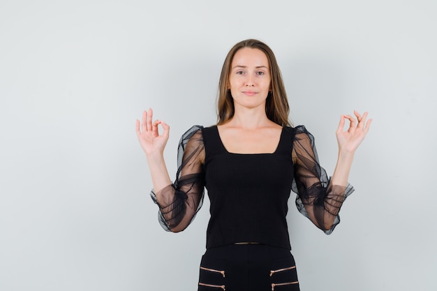 Jovem mulher mostrando sinal de tudo bem com ambas as mãos na blusa preta e calça preta e parecendo otimista. vista frontal.