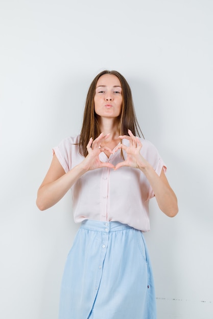Jovem mulher mostrando um gesto de coração com os lábios dobrados em t-shirt, saia, vista frontal.