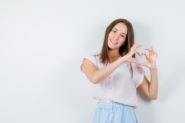 Jovem mulher mostrando um gesto de coração e sorrindo em t-shirt, saia vista frontal.