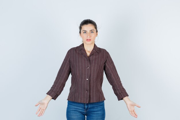 Jovem mulher mostrando um gesto desamparado em camisa listrada, jeans e parecendo perplexo, vista frontal.