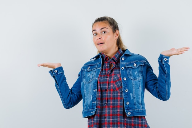 Jovem mulher mostrando um gesto desamparado na camisa, jaqueta e parecendo confusa, vista frontal.