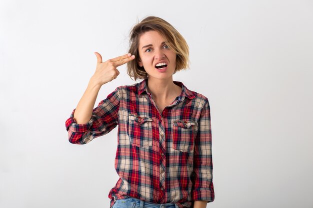 Jovem mulher muito engraçada e emocional com camisa quadriculada posando isolada na parede branca do estúdio, mostrando o gesto da arma