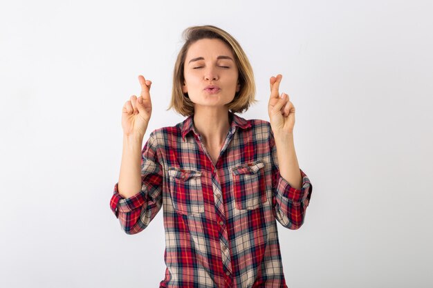 Jovem mulher muito engraçada e emocional com camisa quadriculada posando isolada na parede branca do estúdio, mostrando um gesto de sorte