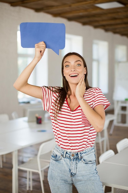 Jovem mulher muito feliz em camiseta listrada olhando espantada na câmera segurando a forma de papel azul da mensagem na mão sob a cabeça no trabalho no escritório vazio moderno