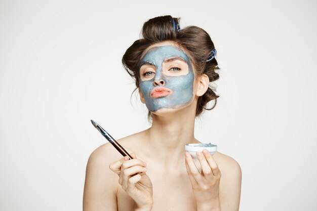 Jovem mulher nua bonita em rolos de cabelo, cobrindo o rosto com mack. Tratamento facial. Cosmetologia de beleza e spa.