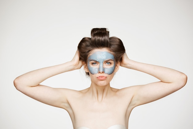 Jovem mulher nua em rolos de cabelo e máscara facial, franzindo a testa. Cuidados com a pele e cosmetologia.
