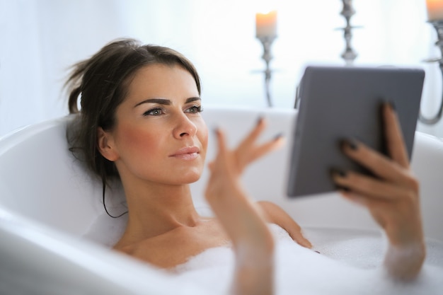 Jovem mulher nua tomando um relaxante bathand espumoso usando tablet digital
