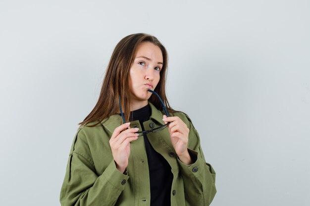 Foto grátis jovem mulher olhando para longe, segurando os óculos na jaqueta verde e olhando pensativa. vista frontal.