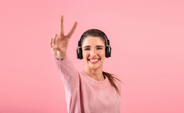 Jovem mulher ouvindo música em fones de ouvido sem fio, vestindo um suéter rosa e sorrindo, posando em rosa