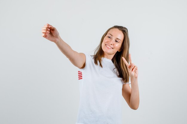 Jovem mulher posando enquanto mostra o sinal de vitória em uma camiseta branca e parece feliz. vista frontal.
