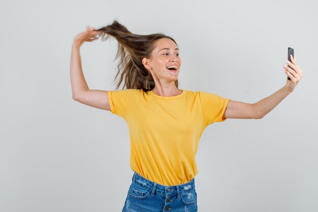 Jovem mulher posando enquanto tira uma selfie de camiseta, shorts e aparência alegre