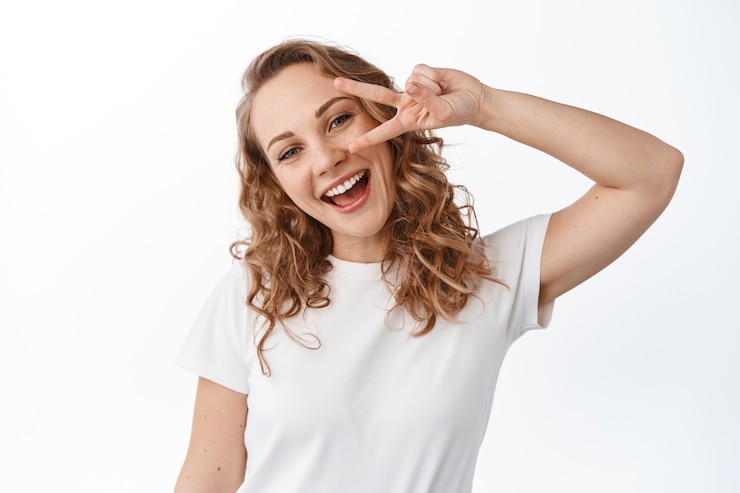 Foto grátis jovem mulher positiva com cabelo loiro encaracolado, maquiagem natural, sorrindo feliz e mostrando o sinal v, gesto de paz, em pé sobre uma parede branca