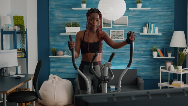 Jovem mulher praticando esportes aeróbicos em bicicleta elíptica, assistindo a um vídeo aeróbico na TV durante a rotina matinal de peso corporal na sala de estar