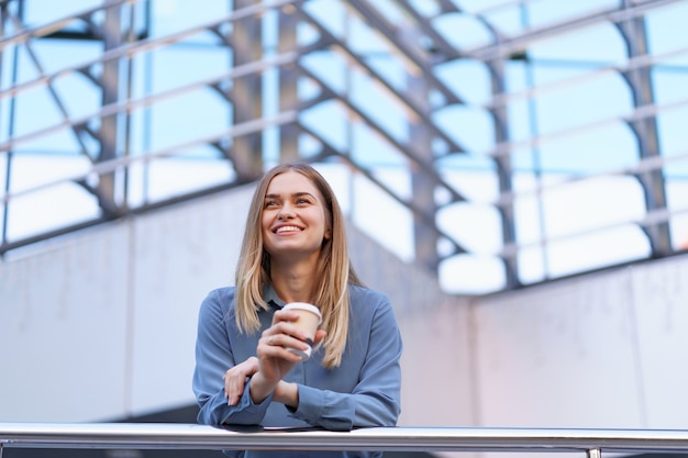 Jovem mulher profissional sorridente, fazendo uma pausa para o café durante seu dia inteiro de trabalho. ela segura um copo de papel ao ar livre perto do prédio comercial, enquanto relaxa e aprecia sua bebida.