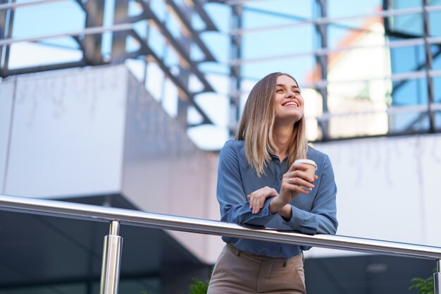 Jovem mulher profissional sorridente, fazendo uma pausa para o café durante seu dia inteiro de trabalho. Ela segura um copo de papel ao ar livre perto do prédio comercial, enquanto relaxa e aprecia sua bebida.