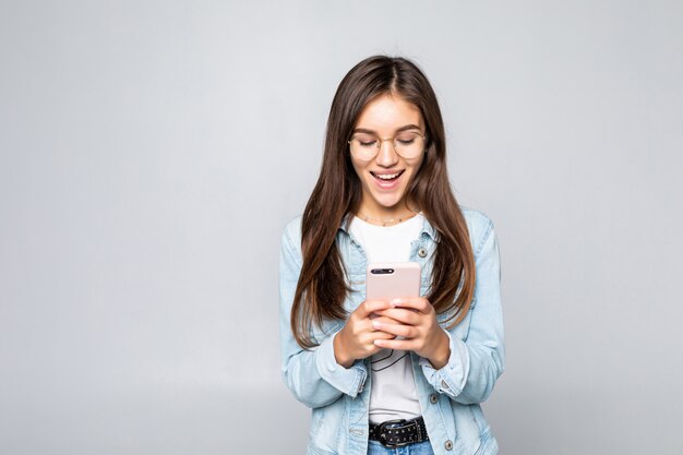 Jovem mulher que sorri e que texting em seu telefone móvel, isolado sobre a parede branca.