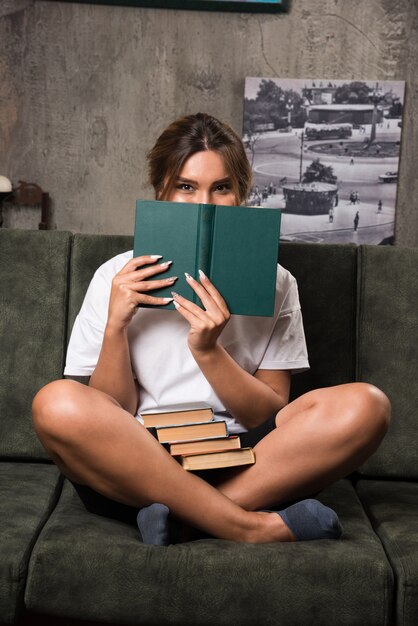 Jovem mulher se escondendo atrás de um livro no sofá.