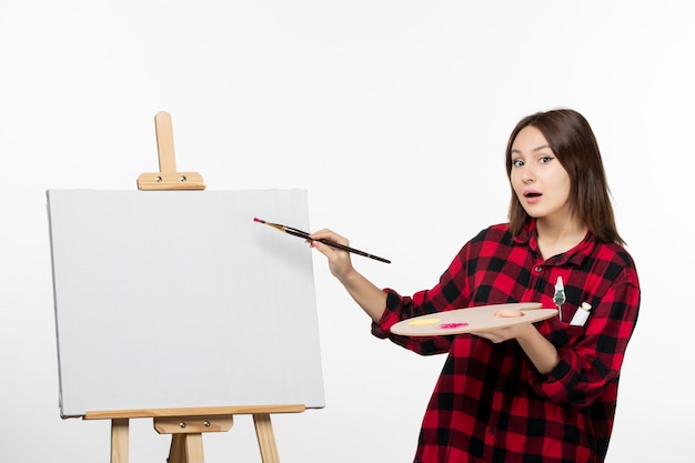 Jovem mulher se preparando para desenhar com pincel em cavalete na parede branca, pintura de exposição de arte com borla do artista, vista frontal