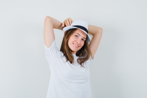 Jovem mulher segurando as mãos levantadas atrás da cabeça em camiseta branca, chapéu e parecendo relaxado.