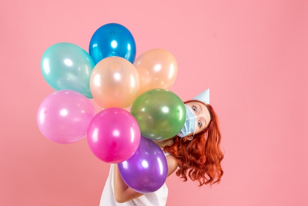 Foto grátis jovem mulher segurando balões coloridos na máscara de frente para o rosa claro