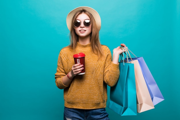 Jovem mulher segurando café para ir e sacolas de compras enquanto sorrindo na parede azul