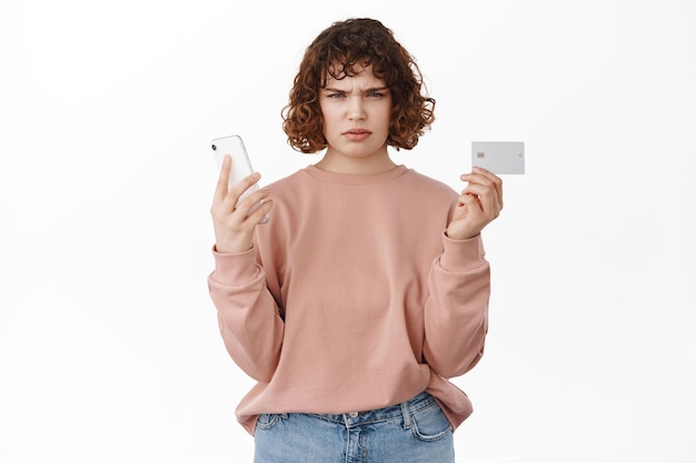 Jovem mulher segurando cartão de crédito e telefone celular, mostrando expressão de rosto zangado, fundo branco