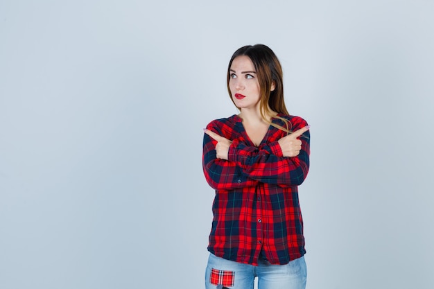 Jovem mulher segurando dois braços cruzados, apontando para direções opostas com os dedos indicadores em uma camisa xadrez, jeans e parecendo indecisa. vista frontal.