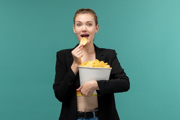 Jovem mulher segurando e comendo batatinhas de frente, assistindo filme na superfície azul