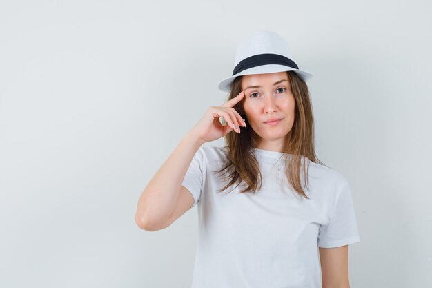 Jovem mulher segurando o dedo nas têmporas em t-shirt branca, chapéu e aparência sensata.