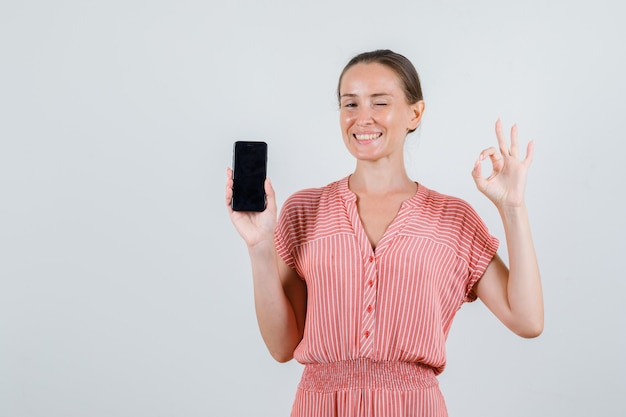 Jovem mulher segurando o telefone móvel com sinal ok em vestido listrado e parecendo alegre. vista frontal.