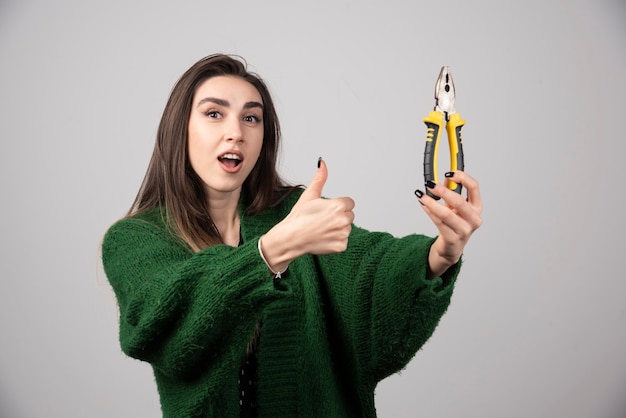 Jovem mulher segurando um alicate e aparecendo um polegar.