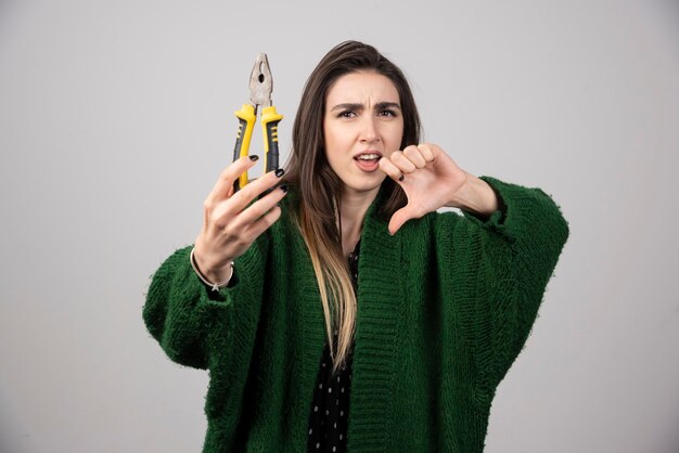 Jovem mulher segurando um alicate e mostrando um polegar para baixo.