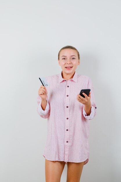 Jovem mulher segurando um celular e um cartão de plástico em uma camisa rosa e parecendo feliz