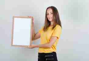 Foto grátis jovem mulher segurando um quadro branco e sorrindo com uma camiseta amarela