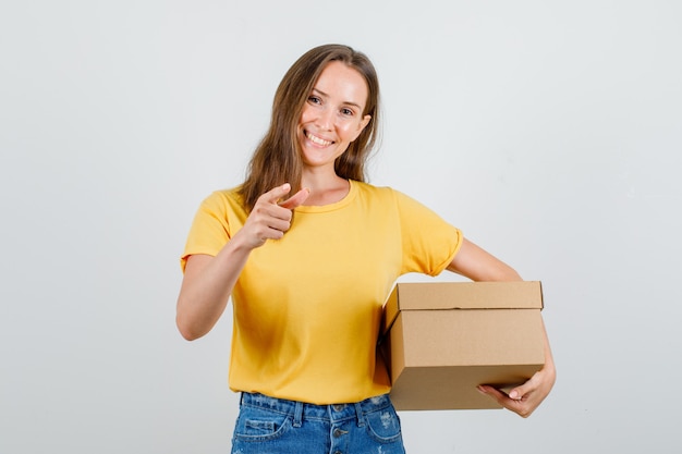 Jovem mulher segurando uma caixa de papelão com o dedo fazendo o sinal em uma camiseta, shorts e parecendo feliz