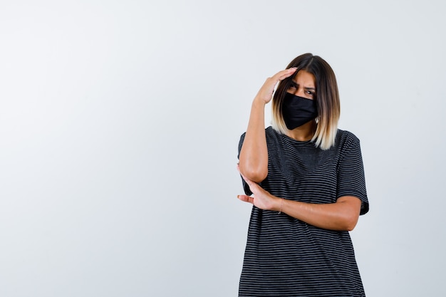 Jovem mulher segurando uma mão na testa, a outra mão sob o cotovelo no vestido preto, máscara preta e olhando sério, vista frontal.