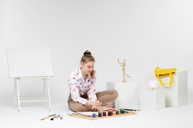 Jovem mulher sentada de frente com tintas e cavalete para desenhar na mesa branca arte desenhar mulher pintor cavalete cor do artista