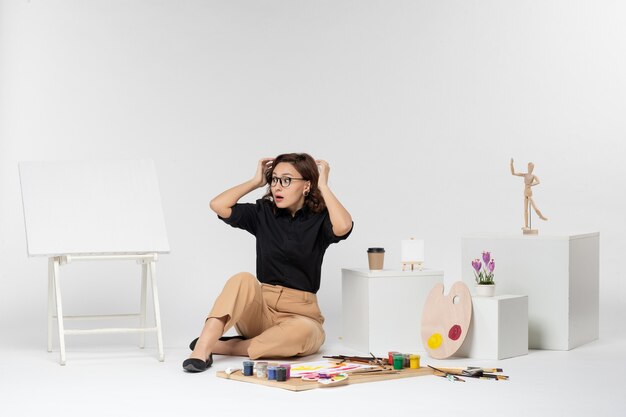 Jovem mulher sentada de frente na sala com cavalete e tintas em fundo branco
