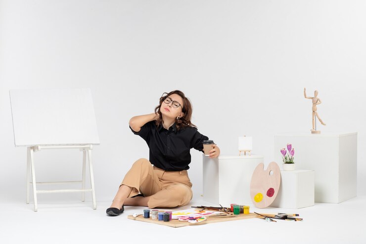 Foto grátis jovem mulher sentada de frente para a sala com tintas e cavalete, com dor de pescoço no fundo branco