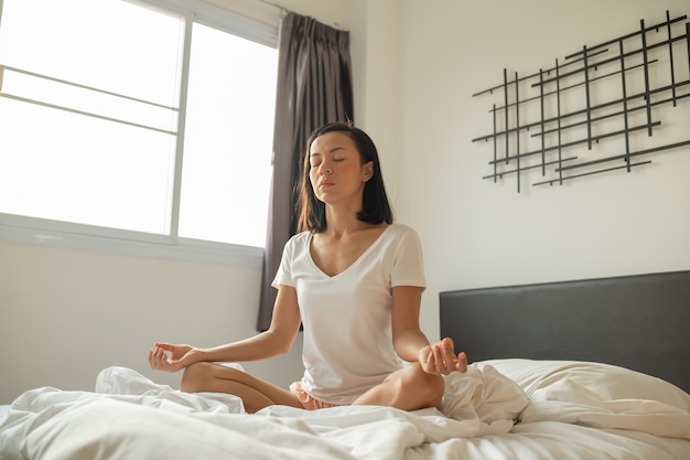 Jovem mulher sentada em sua cama no quarto e meditando.