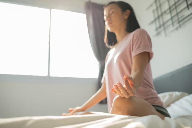 Jovem mulher sentada em sua cama no quarto e meditando.