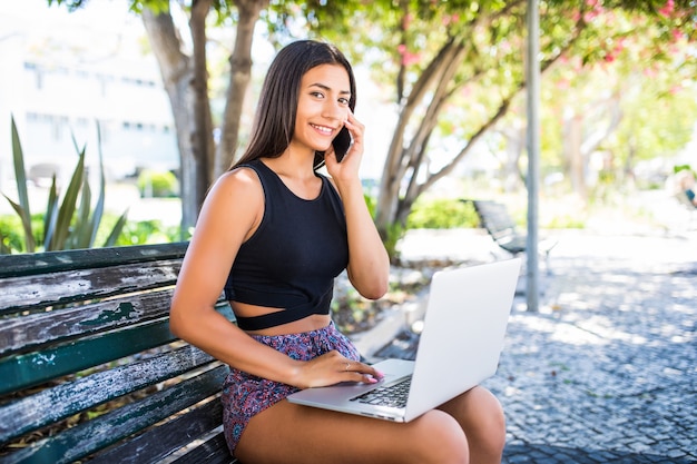 Jovem mulher sentada no banco, falando no smartphone, trabalhando no laptop ao ar livre.