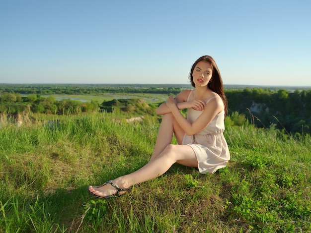 Jovem mulher sentada perto de um penhasco ao ar livre na natureza. Garota atraente com um vestido branco posando ao ar livre. Modelo feminino posando em um campo em um dia ensolarado de verão.